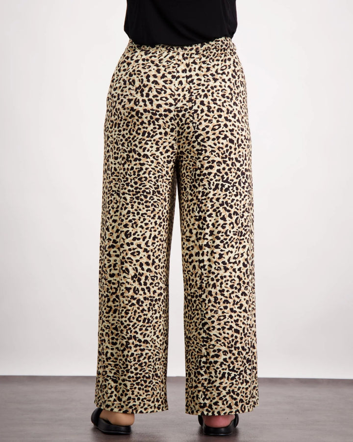 Image is of a female wearing a pair of leopard print bamboo wide leg pants and black leather slippers. She is facing towards the back of the room. The back elastic gather is visible on the pants. The bottom of her black top is visible. Christina Stephens Australian Adaptive Clothing.