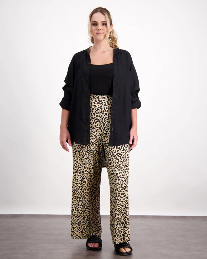 A standing female with caucasian skin and sandly coloured curly hair is wearing a black camisole under a black open linen shirt. She is wearing leopard print wide leg pants and black leather slippers. Christina Stephens Australian Adaptive Clothing. 
