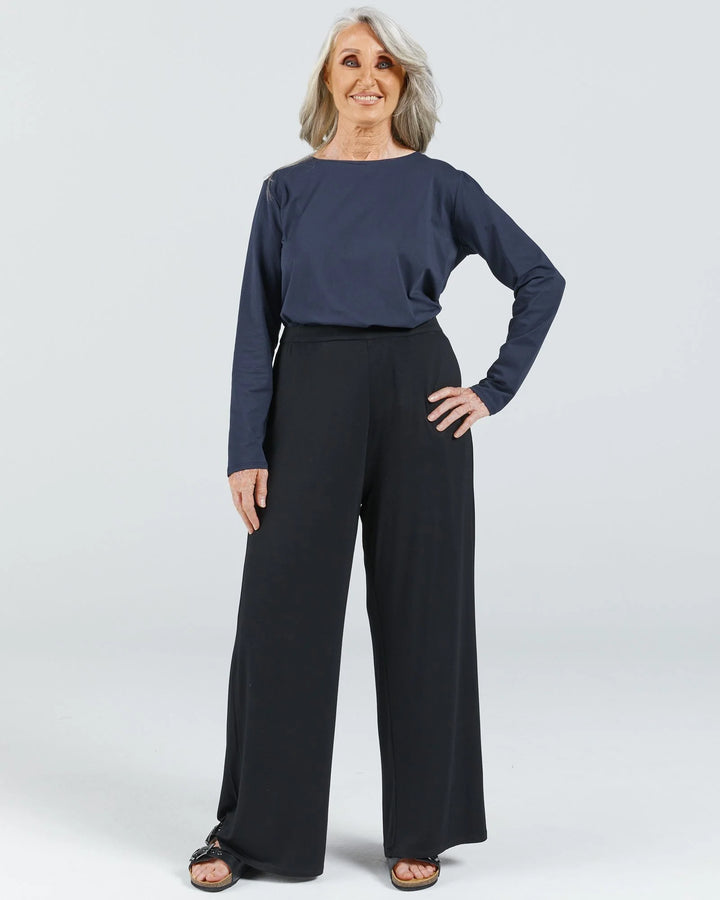 Image is of a female wearing a pair of black bamboo wide leg pants, a navy long sleep leaf-back t-shirt and black leather slippers.  She has grey hair and her hand is resting on her hip. Christina Stephens Australian Adaptive Clothing.